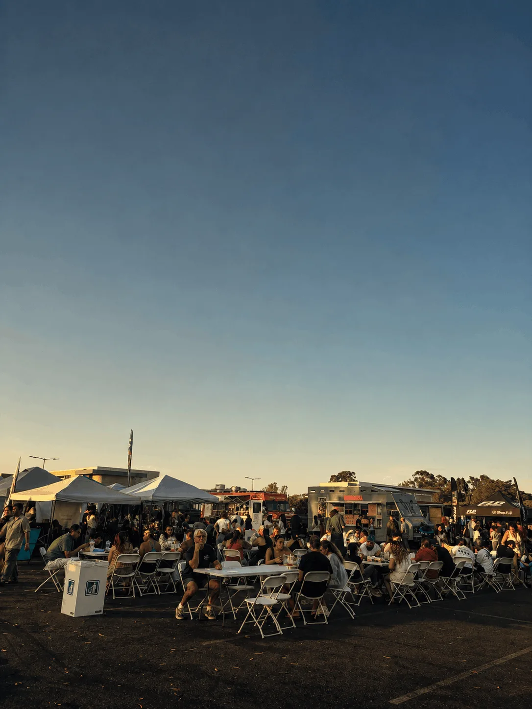 Gente comiendo en mesas largas y compartidas frente a food trucks y carpas de vendedores al atardecer, Long Beach Foodies Market